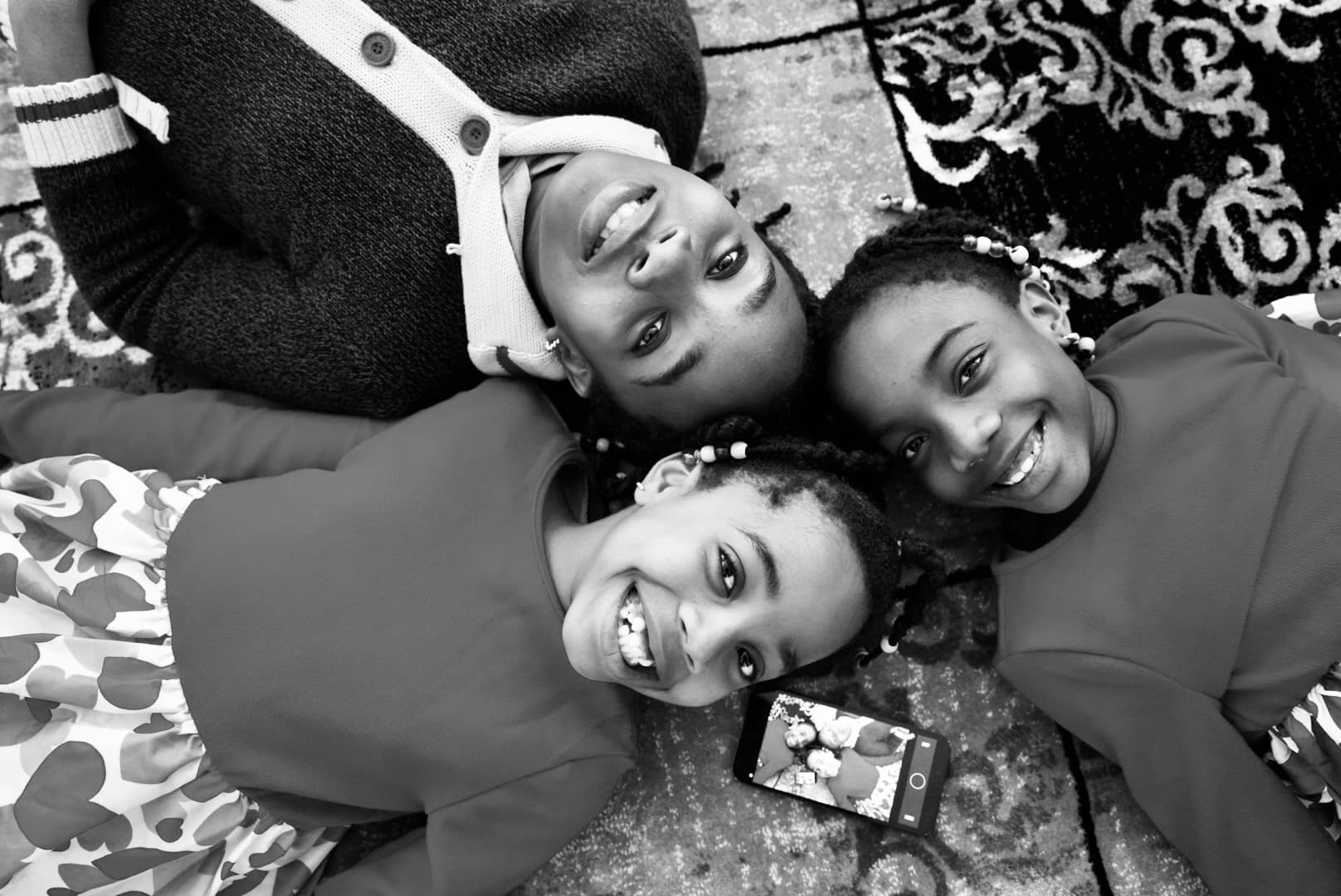 Black and white photograph of three young girls lying on a patterned rug, heads together, smiling up at the camera